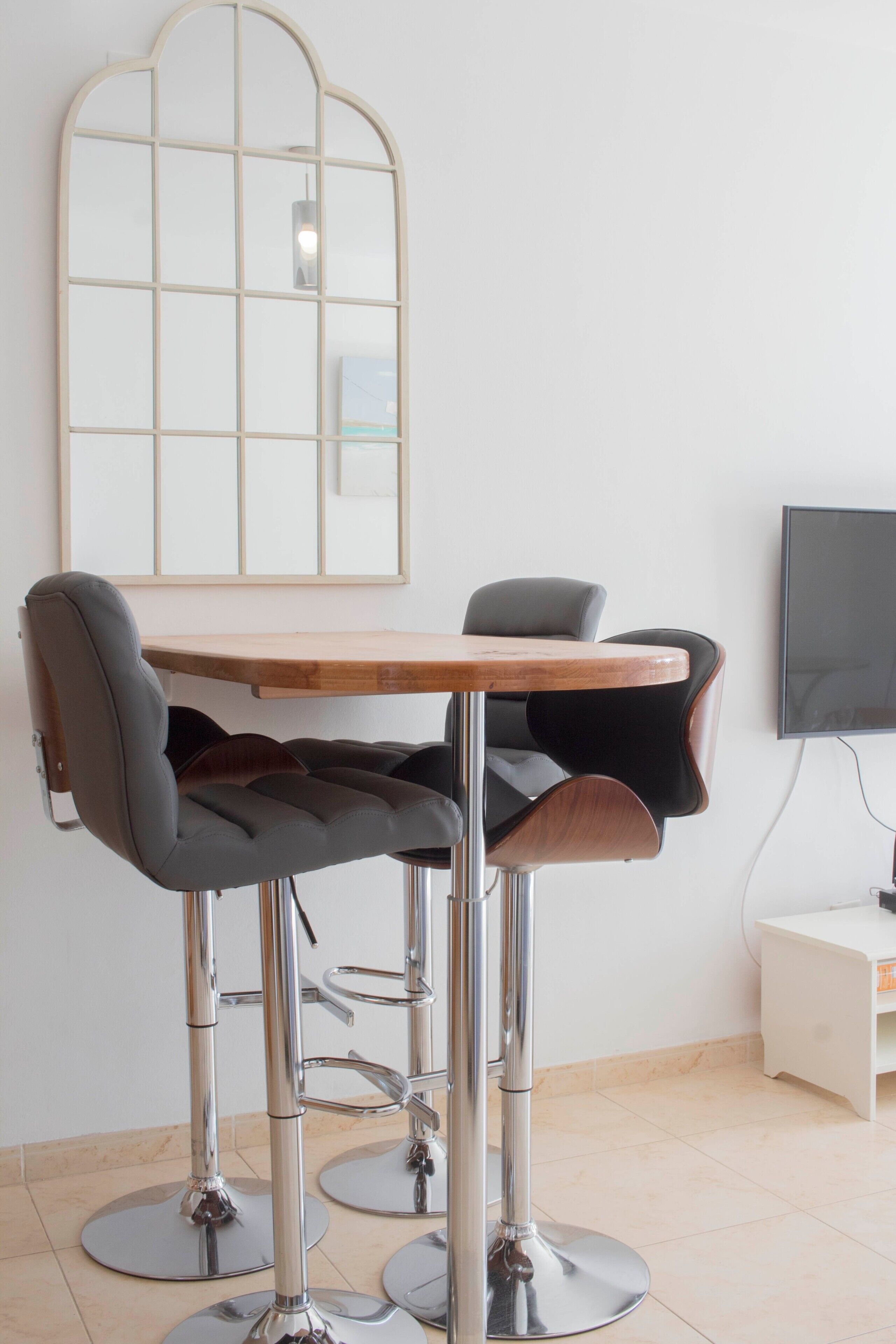 Un espace bar moderne avec une table en bois et deux tabourets de bar, un grand miroir et un téléviseur à écran plat fixé au mur.