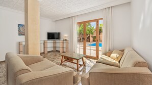 Un salon avec des canapés de couleur beige, une table basse en verre, un téléviseur et une vue sur une piscine.