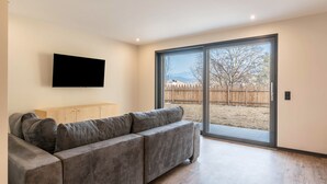 Een moderne woonkamer met een grijze modulaire bank, een flatscreen televisie aan de muur en een schuifpui die leidt naar een buitenruimte.