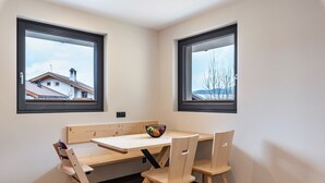 Una habitación de esquina con una mesa y sillas de madera, dos ventanas con marcos negros y un frutero sobre la mesa.
