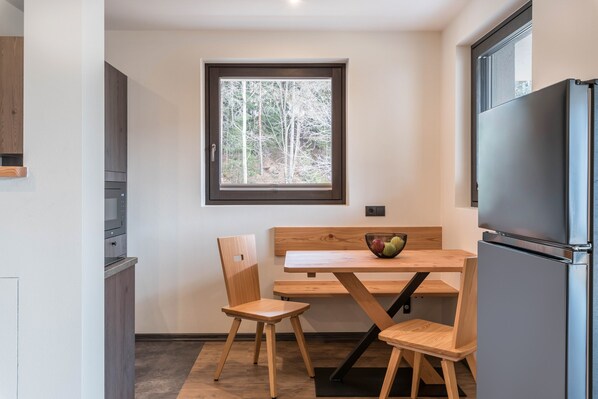 Une cuisine de taille réduite, avec une table en bois, deux chaises, un réfrigérateur et une fenêtre donnant sur des arbres.