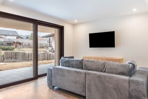 Un salon moderne avec un canapé gris, un téléviseur à écran plat fixé au mur et une porte-fenêtre coulissante donnant sur un espace extérieur.