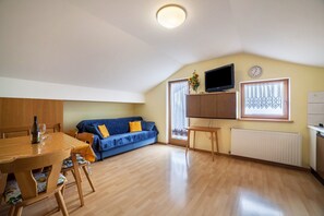 Huoneessa on ruokapöytä, sohva, seinälle asennettu televisio ja ikkuna, jossa on kuviolliset verhot.