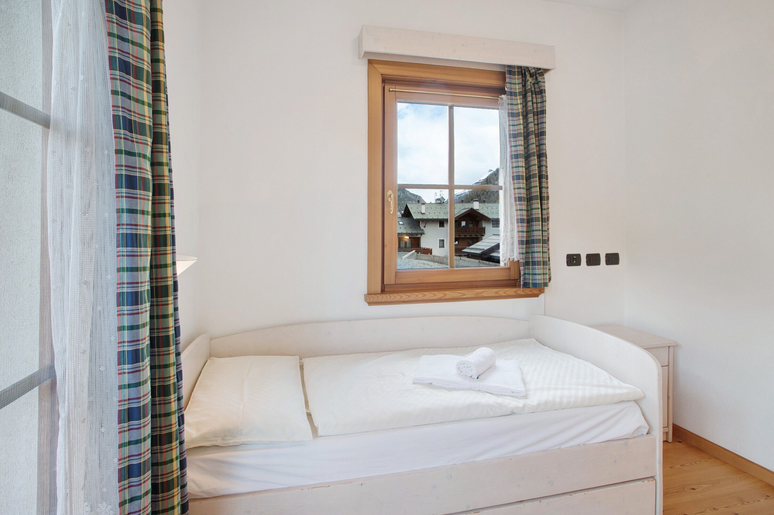 A small, single-bed room with a window, a view of buildings outside, and a wooden headboard.