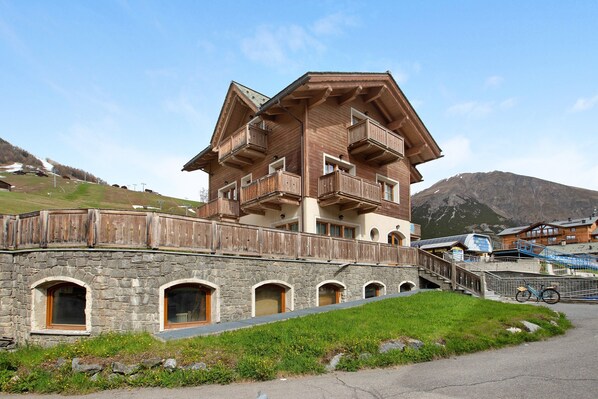 A multi-story wooden building with balconies, stone lower level, and a mountain backdrop.