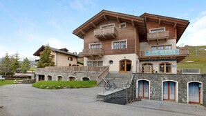 Un immeuble à plusieurs étages, avec une enseigne indiquant un service de location de matériel de ski, des marches en pierre et un balcon en bois.
