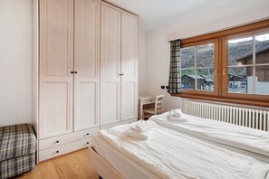 Une chambre dotée d’une grande armoire, d’un lit avec du linge de maison blanc, d’un petit bureau et d’une fenêtre donnant sur des bâtiments et des collines.