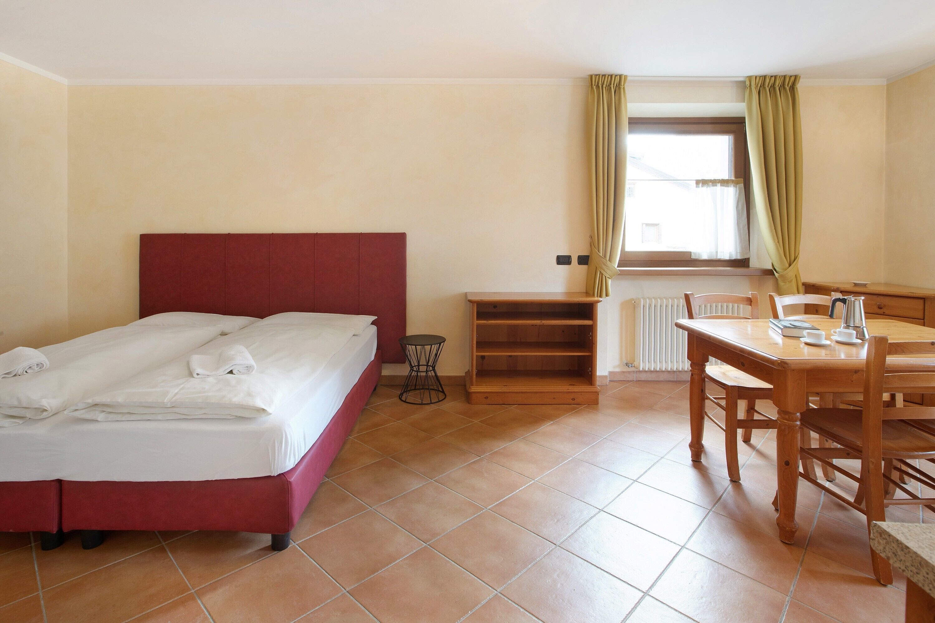 Habitación de hotel con cama, una mesita, una silla, un radiador y una ventana con cortinas.