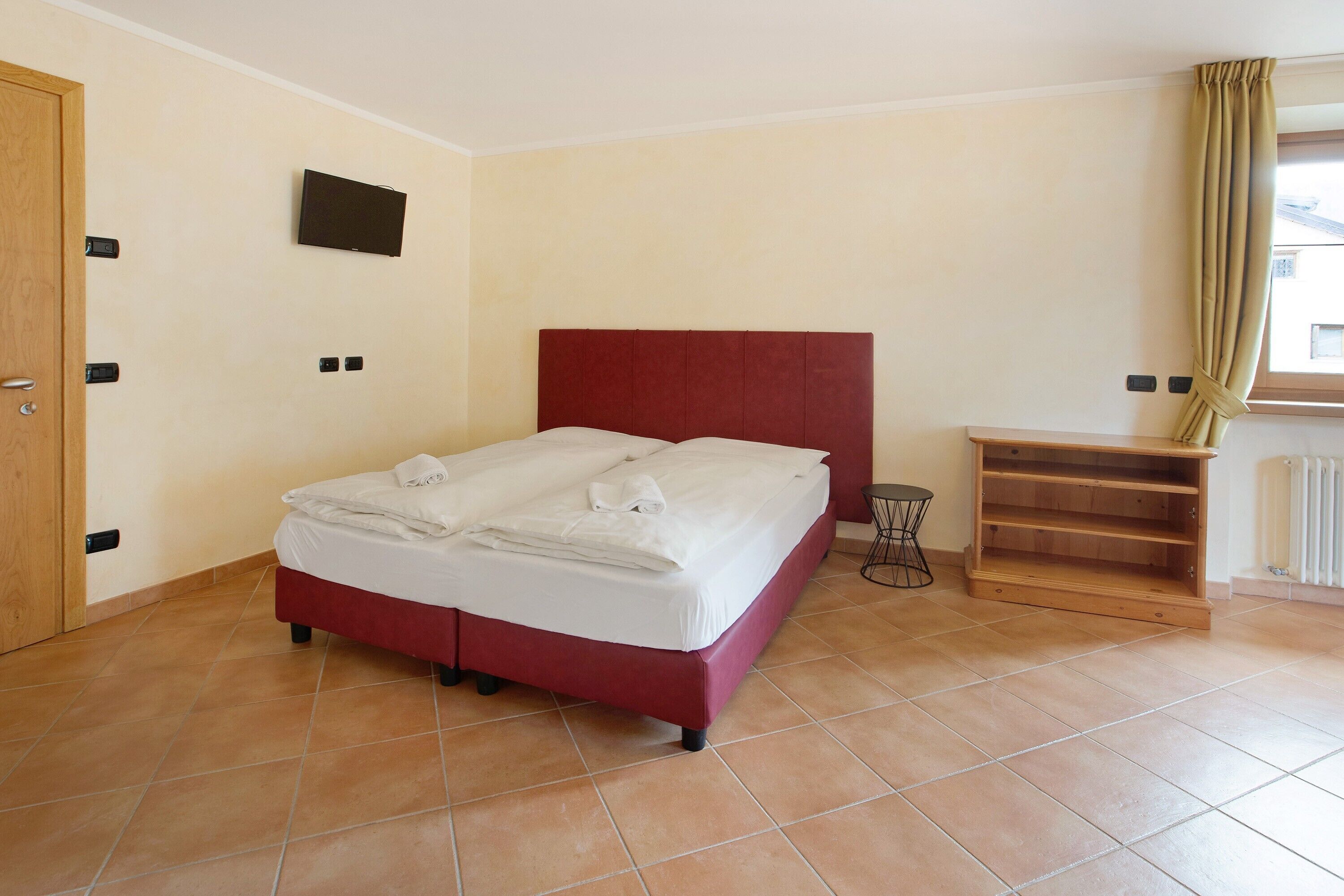 Habitación de hotel con cama, televisión montada en la pared, radiador y ventana con cortinas.