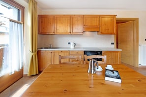 A kitchen with wooden cabinets, a dining table, chairs, a kettle, a book, and a window with curtains.