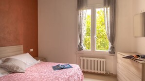 Une chambre à coucher avec un lit, une fenêtre avec des rideaux, un radiateur et une commode sur laquelle se trouve un magazine.