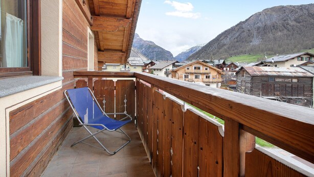 Ein Balkon mit Holzgeländer, einem blauen Klappstuhl und Blick auf ein Bergdorf.