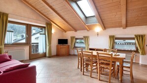 Ein Zimmer mit einer Couch, einem Esstisch, Stühlen, einem Fernseher und Fenstern mit Vorhängen.