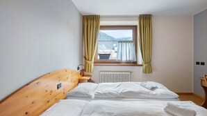 Ein Schlafzimmer mit zwei Betten, einem Holzkopfende, einem Fenster mit Vorhängen, einem Heizkörper und Blick auf Berge.