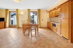 Une cuisine avec des meubles en bois, une table à manger avec des chaises, un téléviseur et une vue sur l’extérieur, visible à travers une porte.