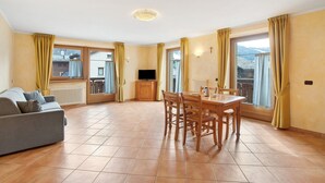 Ein geräumiger Wohnbereich mit einer Couch, einem Esstisch, Stühlen, großen Fenstern mit Vorhängen und einem Fernseher auf einem Schrank.