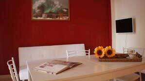 A dining area with a red wall, a painting, a television, and a table with a magazine, sunflowers, and a tray. - Apartment 'Il Fosso' with Sea View, Wi-Fi (Borghetto Santo Spirito)