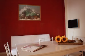 Un comedor con pared roja, un cuadro, un televisor y una mesa con una revista, girasoles y una bandeja.