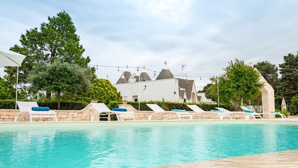 Una piscina con sillas de descanso, un muro de piedra y un edificio con techos cónicos al fondo.
