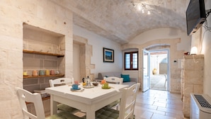 Un comedor con mesa y sillas blancas, una estantería con utensilios de cocina, un cuadro en la pared y un acceso abierto al exterior.