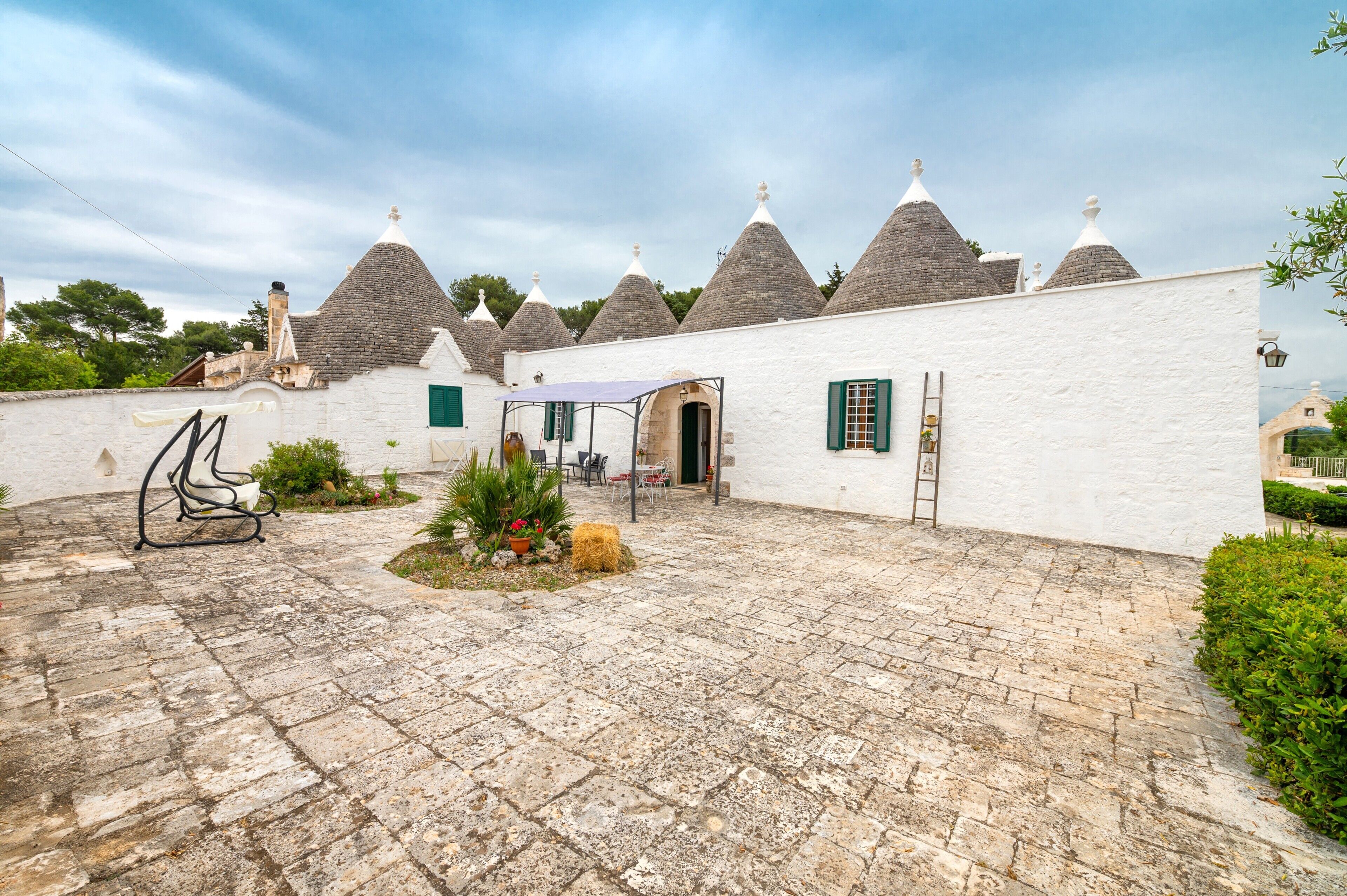A courtyard with stone paving, a white building with conical roofs, and a small garden area with plants and a bench.