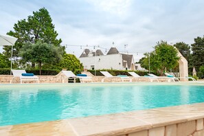 Una piscina con sillas de descanso, un muro de piedra y un edificio con techos cónicos al fondo.