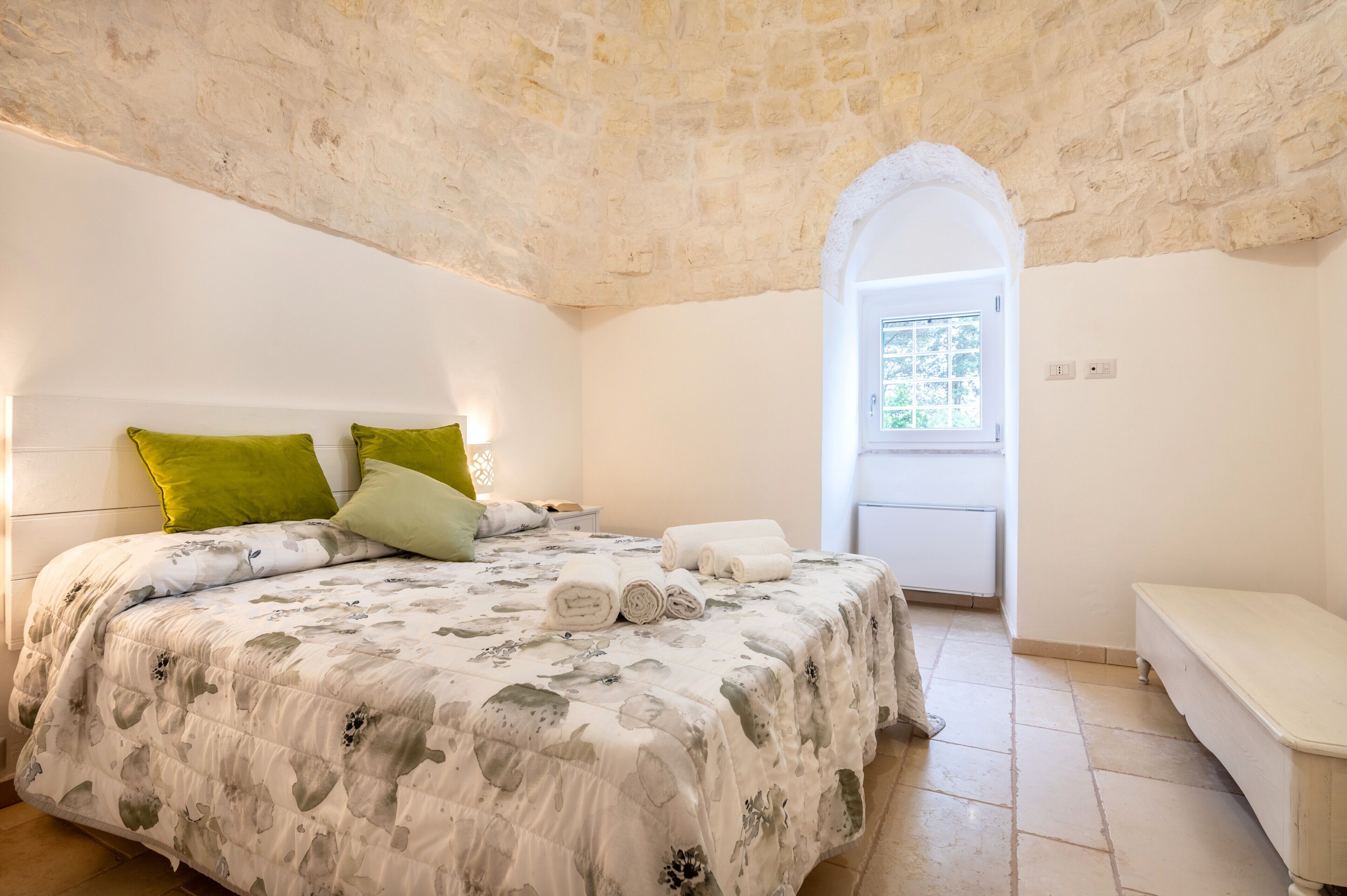 A bedroom with a stone ceiling, a bed with floral bedding, a bench, and a window with a view.