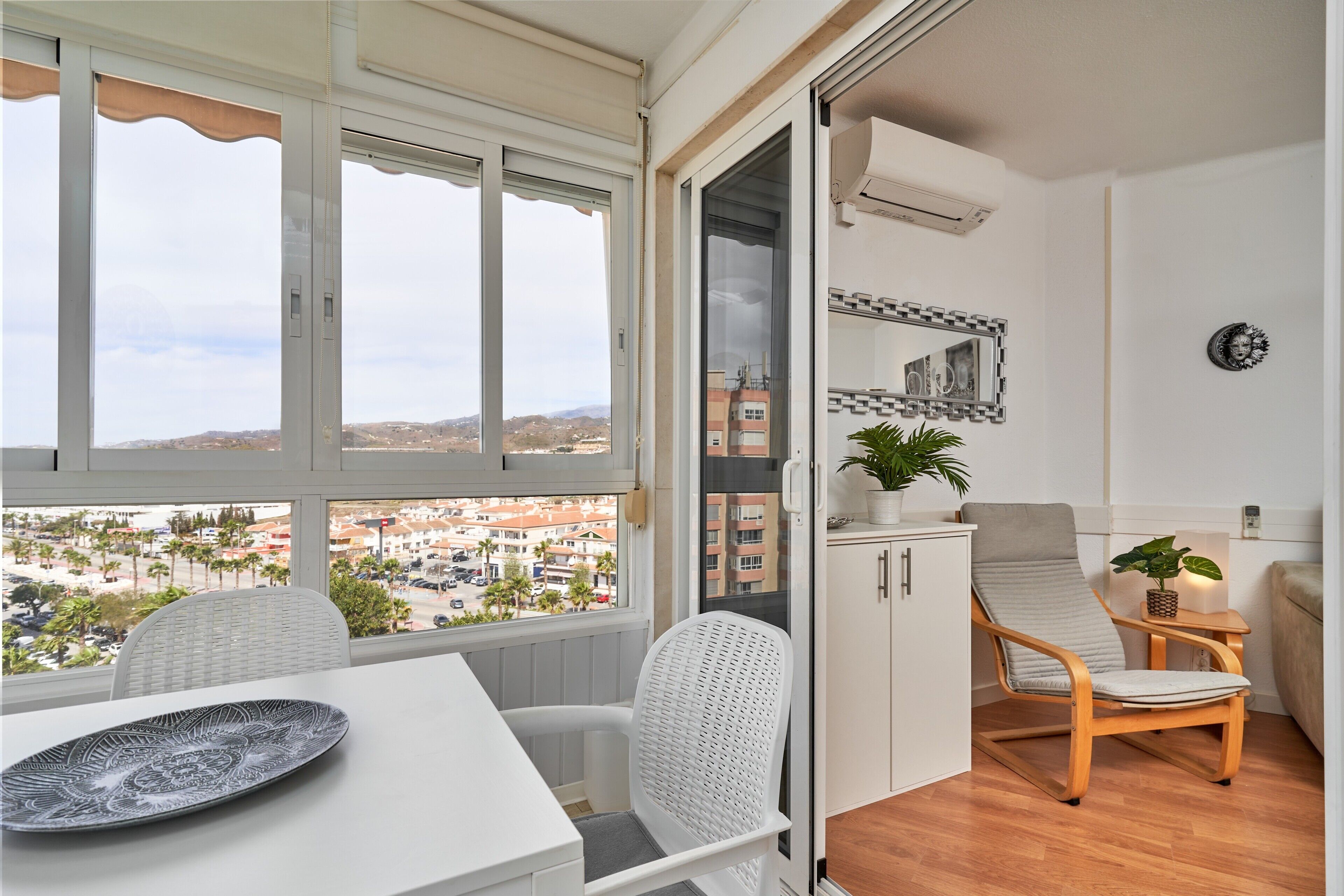 A balcony with a view of a cityscape, a dining table with a plate, a white wicker chair, a wooden chair, and a small cabinet with a plant.
