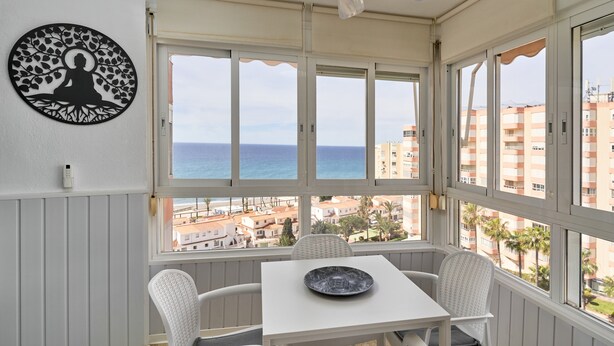 Ein Balkon mit Tisch und Stühlen, einem Deckenventilator und Blick auf das Meer und Gebäude.