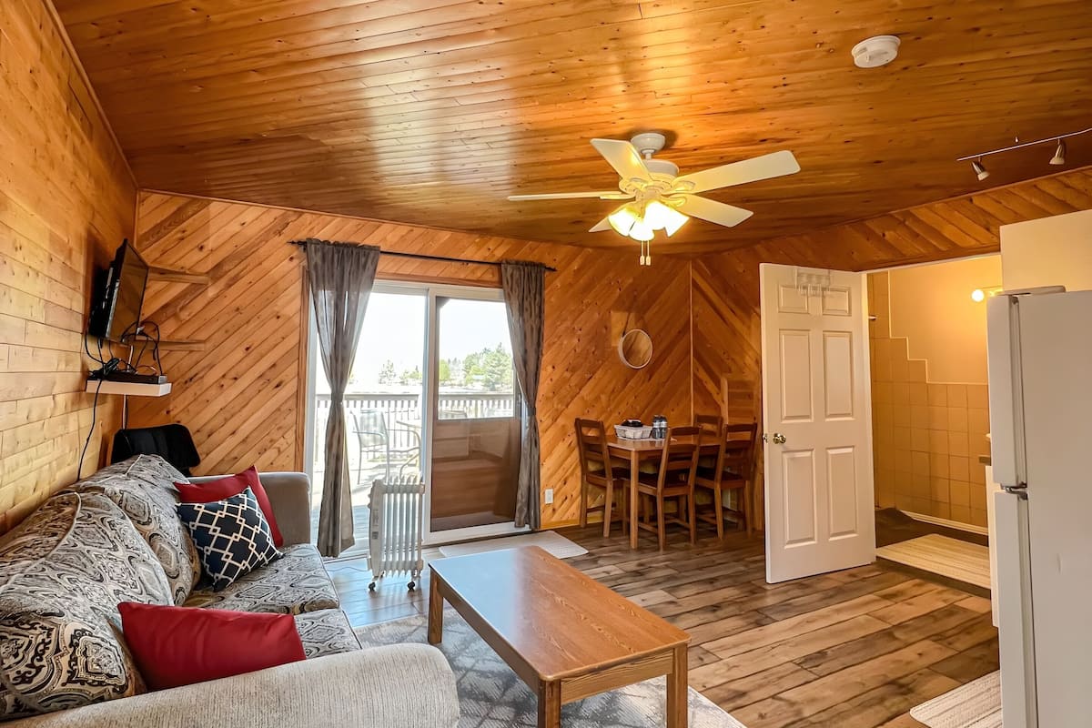 L’intérieur chaleureux d’un chalet avec un plafond en bois, un canapé agrémenté de coussins à motifs, une table à manger et un coin cuisine.
