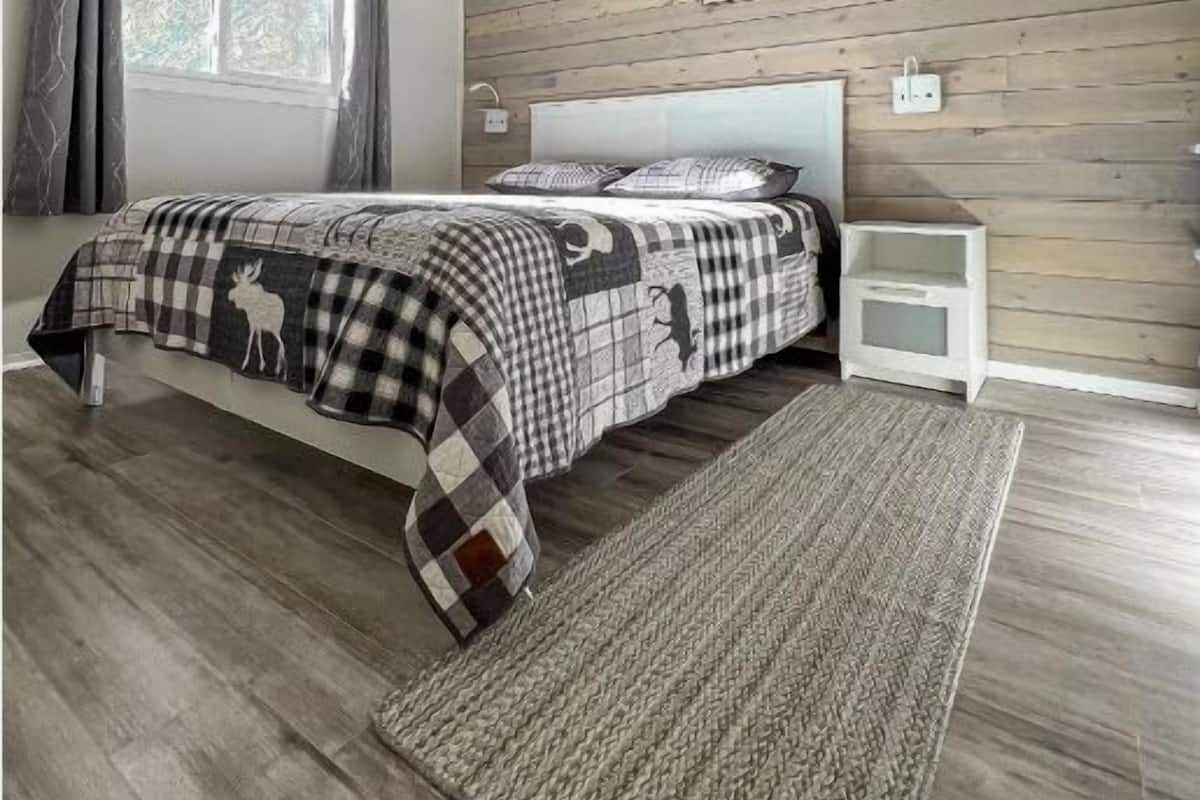 Une chambre avec un plafond en bois, un lit recouvert d’une couverture à carreaux, une table de chevet, une commode et une fenêtre avec des rideaux.