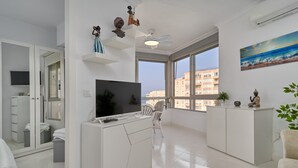 A modern living room with a TV, white furniture, a ceiling fan, large windows, and a view of buildings. - Great Views Studio A84 near the beach with shared pool and Wi-Fi (Torrox)