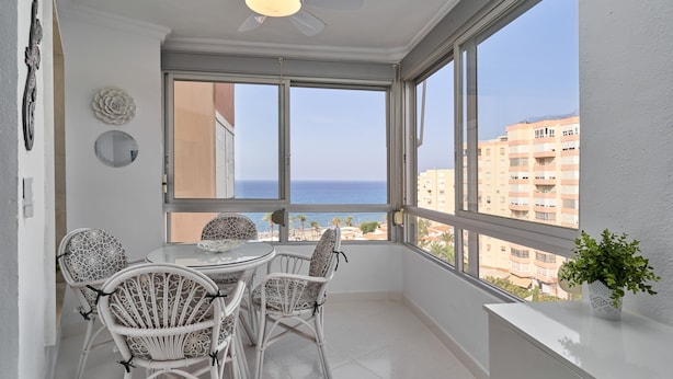 A balcony with a dining table and chairs, large windows with a view of the sea and buildings, and a ceiling fan. - Great Views Studio A84 near the beach with shared pool and Wi-Fi (Torrox)