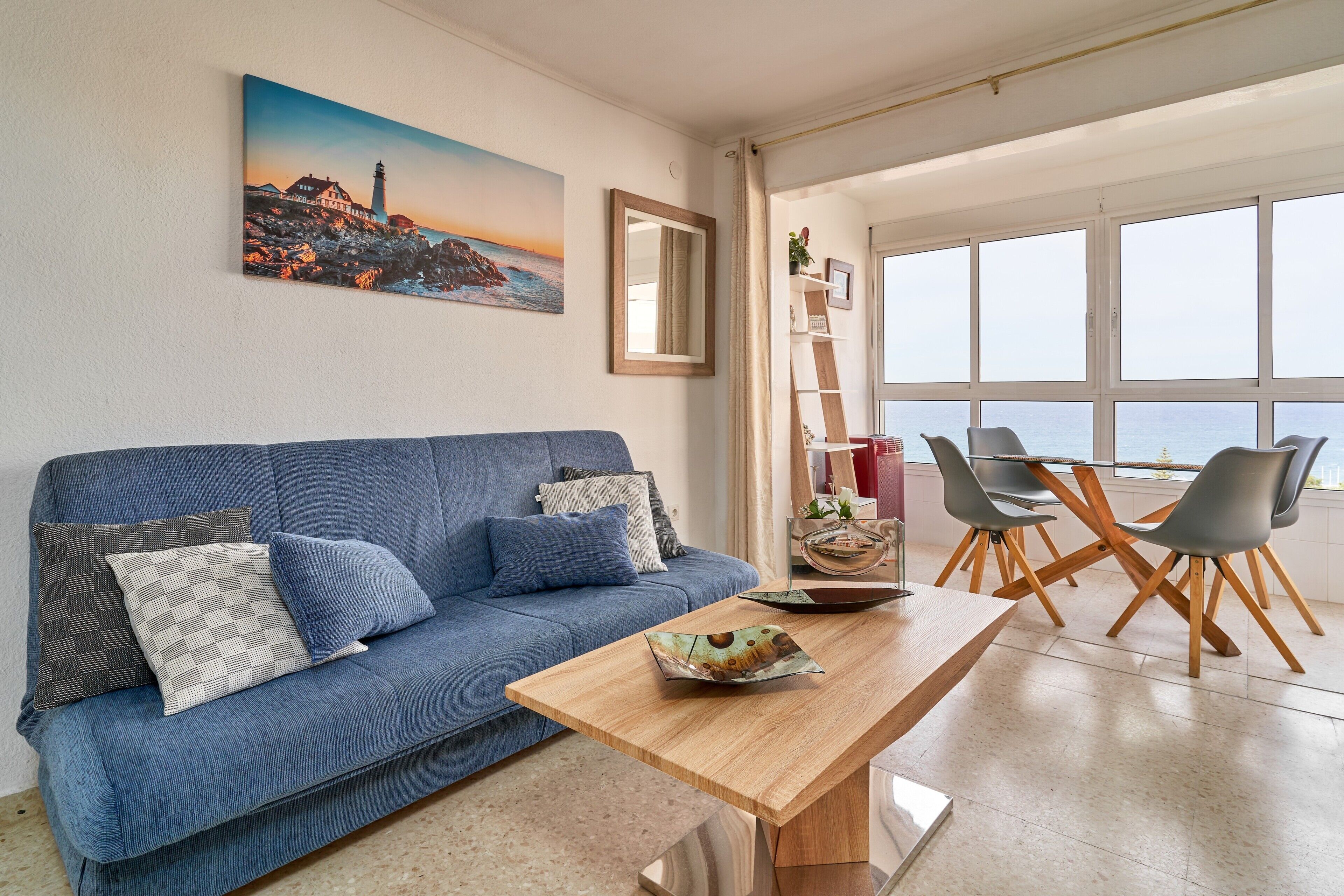 A living room with a blue sofa, a wooden coffee table, and a view of the sea through large windows.