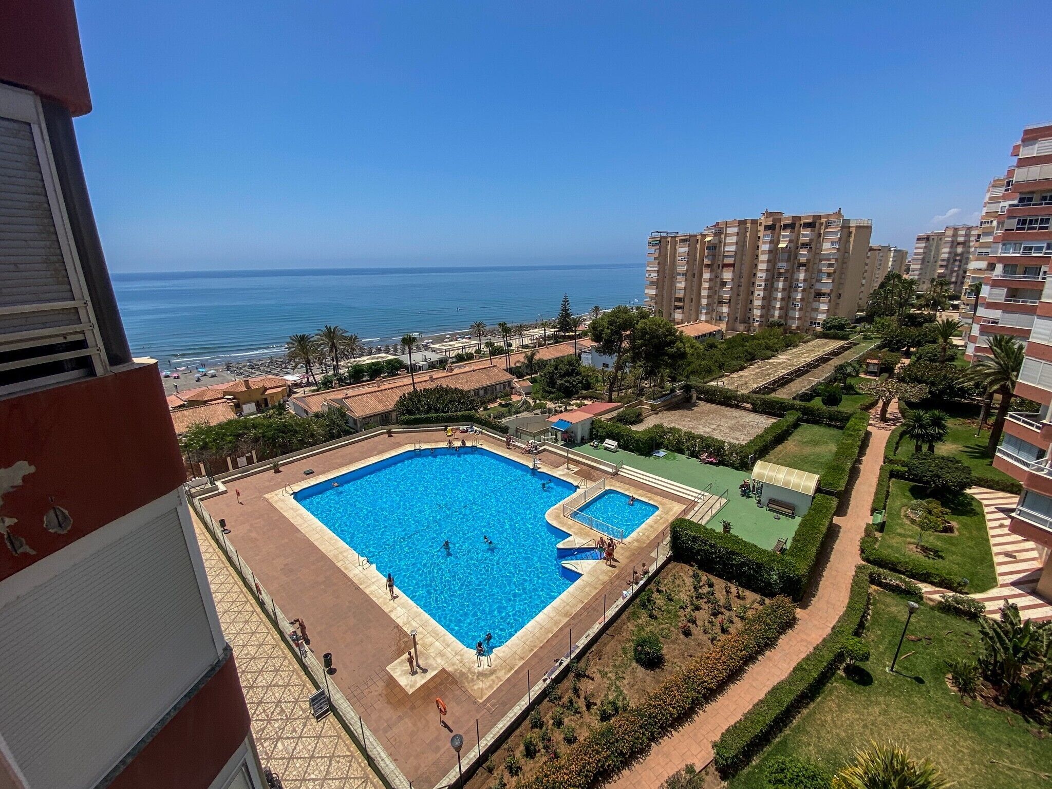 A swimming pool surrounded by greenery, with a view of the sea and high-rise buildings in the background.