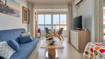 A living room with a sofa, a dining table, chairs, a TV, and a view of the sea.