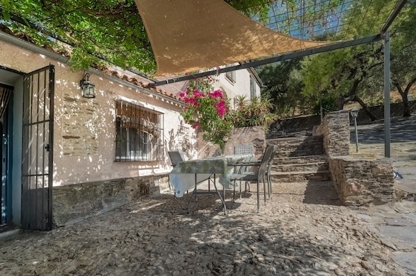 A stone building with a table and chairs under a shade sail, surrounded by greenery and a stone staircase.