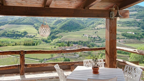A wooden balcony with a table and chairs, overlooking a green valley with fields and houses.