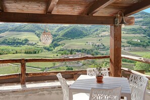 A wooden balcony with a table and chairs, overlooking a green valley with fields and houses. - Cottage Relax – Romantic Retreat Amid Vineyards in Alta Langa – Tenuta MonteOliveto (Vesime)