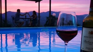 A poolside setting with a glass of red wine and a bottle, a couple dining, and a sunset view.