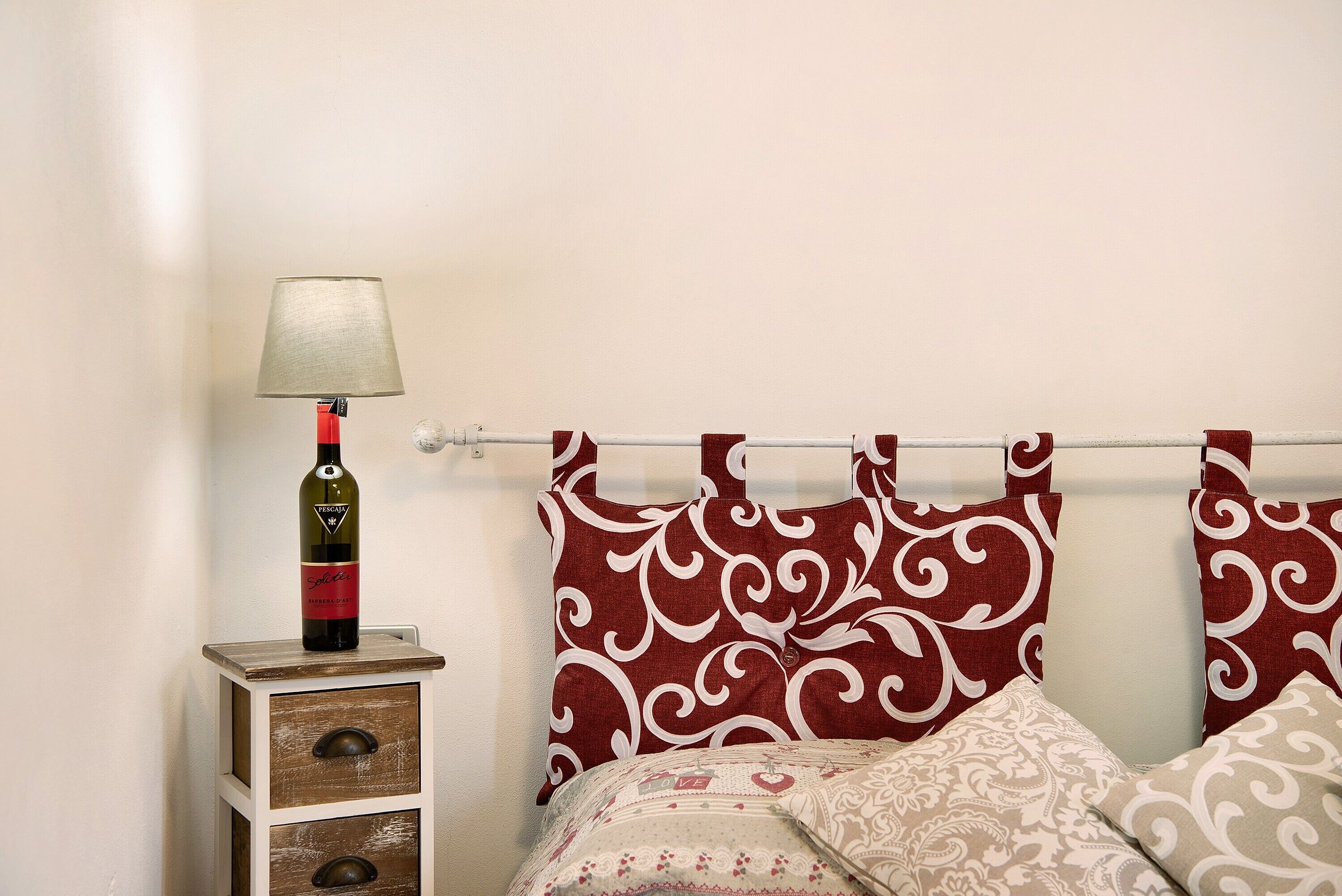 A bed with patterned bedding, a bedside table with a lamp, and a bottle of wine.