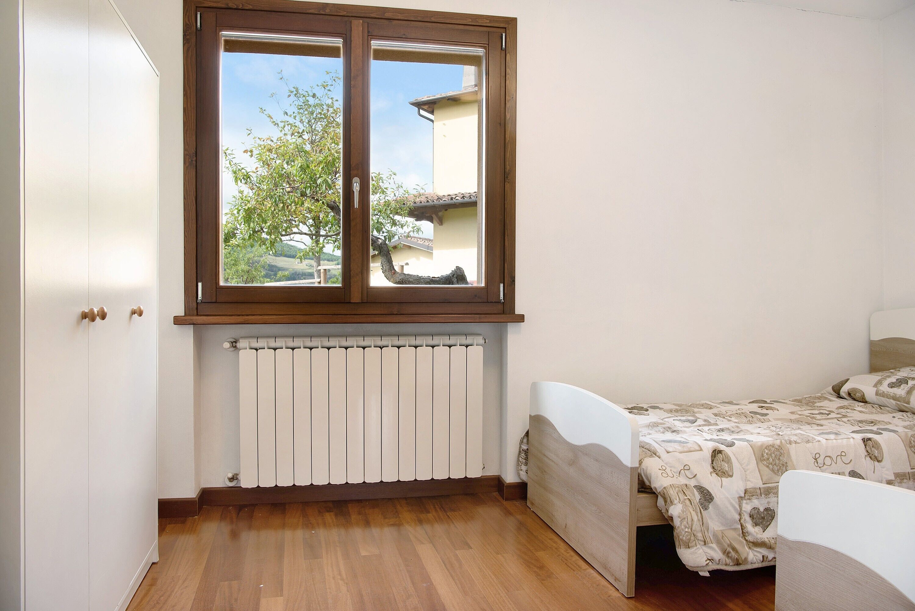 Une chambre avec une fenêtre, un radiateur, un plancher en bois et un lit recouvert d’une couvre-lit à motifs.