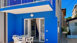 Un edificio azul con balcón, puerta corredera de vidrio que da a una cocina, y un patio con sillas y mesa de plástico blanco.