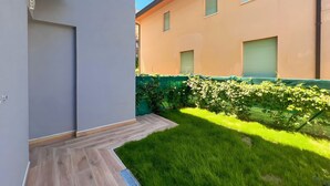 Un edificio moderno con techo plano, un pequeño jardín con terraza de madera y cercado verde.