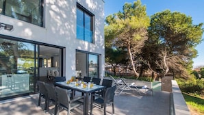 Un espace de repas extérieur moderne, comprenant une table et des chaises, un patio fermé par des baies vitrées, et offrant une vue sur des arbres et un bâtiment.