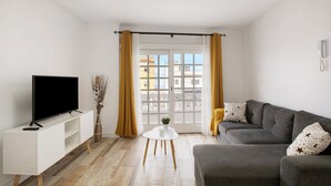 Een woonkamer met een flatscreen televisie op een witte tv-standaard, een grijze bank met gekleurde kussens, een klein wit tafeltje met een plant en een groot raam met gele gordijnen.