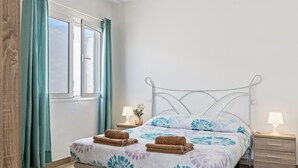 Un dormitorio con cama, dos mesitas de noche, un ventanal con cortinas y una mesita de noche con lámpara.