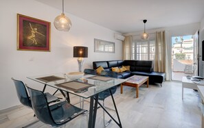 A modern living room with a glass dining table, black leather sofa, and a large framed artwork on the wall.