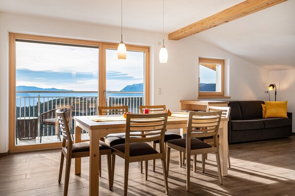 Un comedor con una mesa y sillas de madera, un sofá y un balcón con vistas a las montañas.