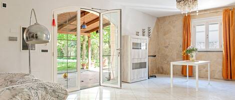 A room with a bed, a white table, a chandelier, a door leading to an outdoor area, and a stone wall.